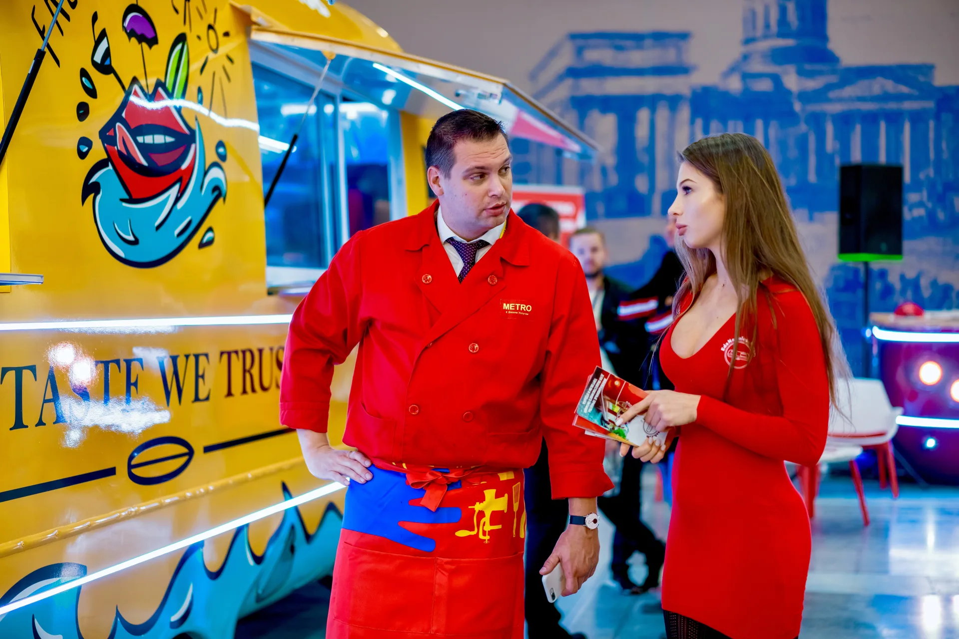 People speaking beside an illustrated yellow mobile food trailer at an indoor event